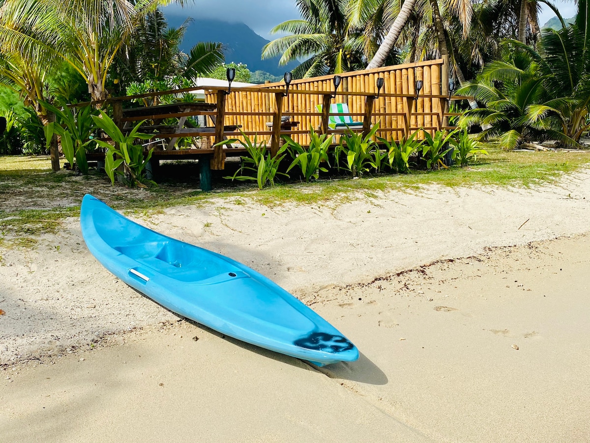 A light blue kayak sits on the sandy beach, close to a wooden deck surrounded by lush tropical plants. The background features palm trees and mountains under a partly cloudy sky, creating a tranquil coastal setting.