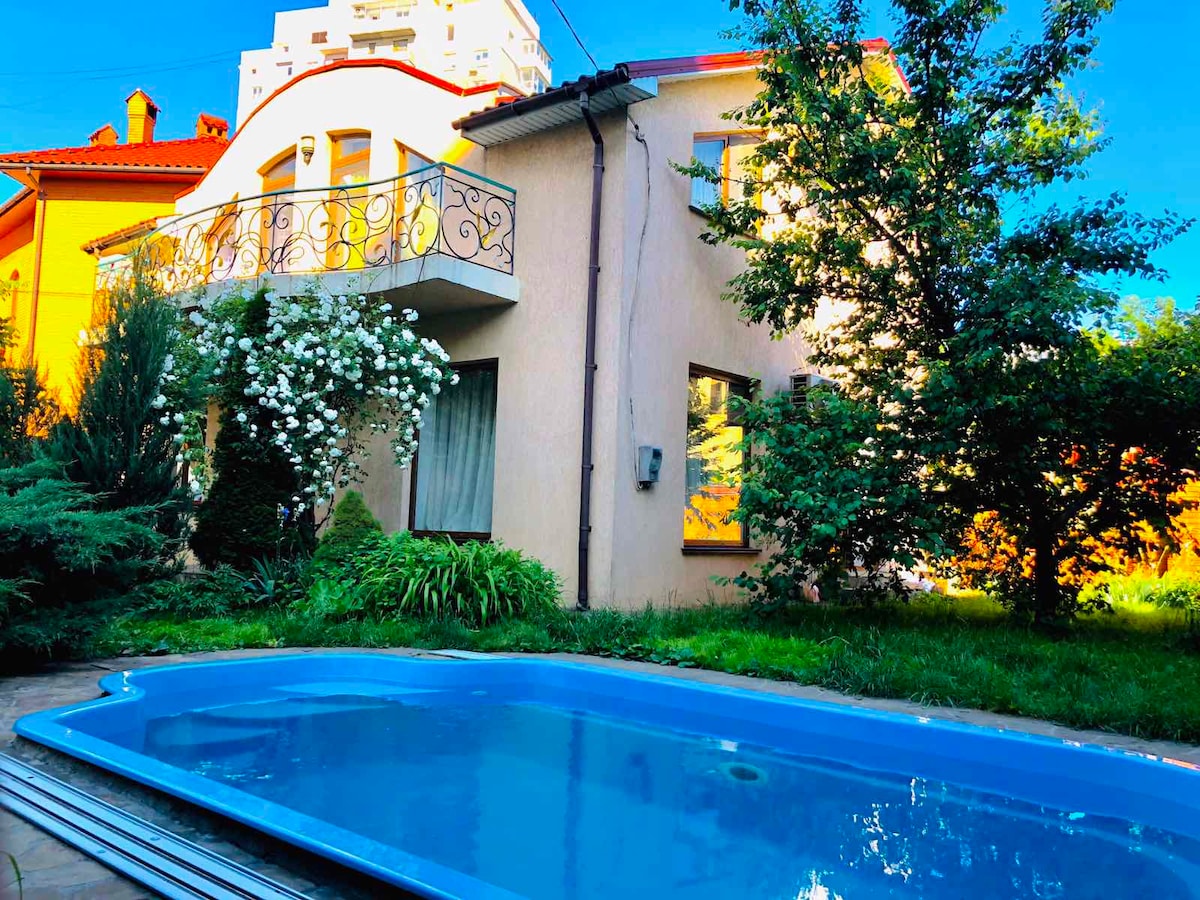 A two-story house is surrounded by greenery and features large windows that allow natural light to fill the interior. A swimming pool is positioned in the foreground, reflecting the blue sky. White flowers add a touch of beauty to the landscape.
