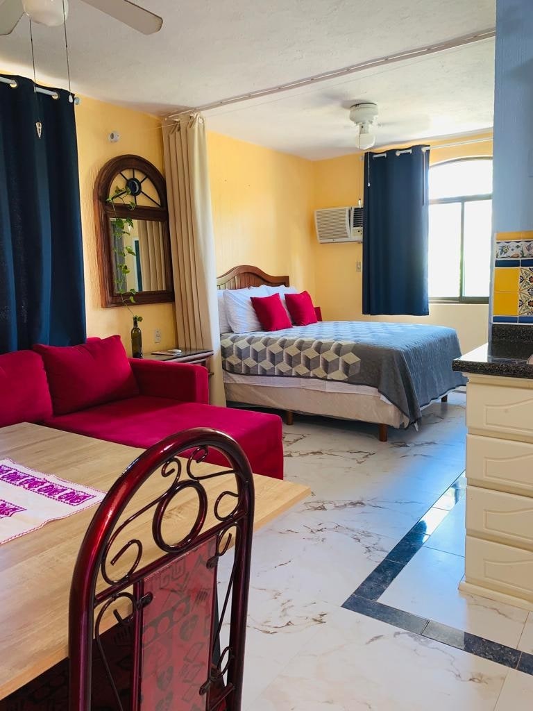 A cozy studio space is shown, featuring a comfortable bed with decorative pillows and a gray quilt. A dining area with a wooden table and red chairs is visible. Light filters through a window with blue curtains, illuminating the cheerful yellow walls.