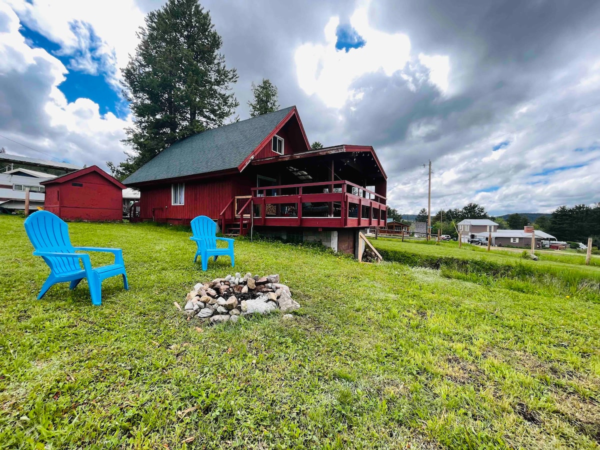A red cabin with a sloped roof is situated on a grassy lot. Bright blue Adirondack chairs are positioned near a small fire pit, surrounded by grass. A large covered deck extends from the cabin, offering outdoor seating and a view of the surrounding area.