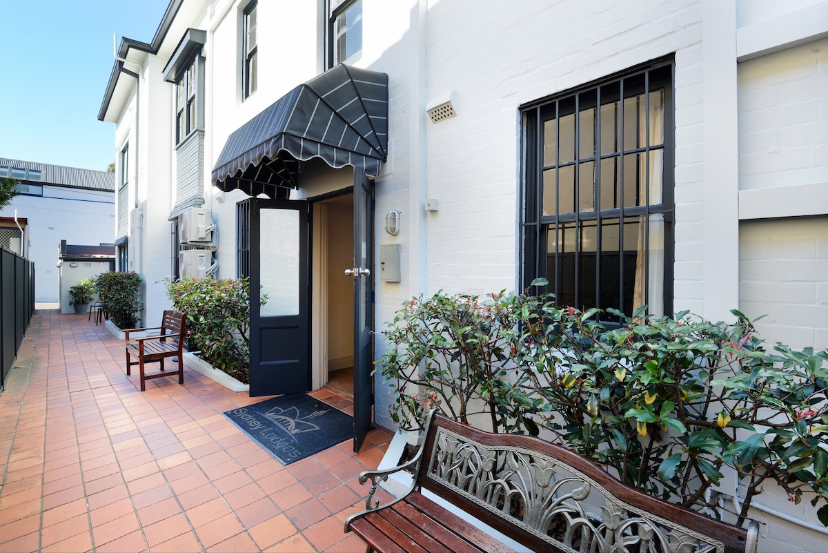An inviting entrance is framed by a stylish awning, displaying a mix of brick and white walls. Lush greenery lines the pathway, which leads to the door. Two wooden benches offer seating along the tiled walkway, enhancing the welcoming exterior.