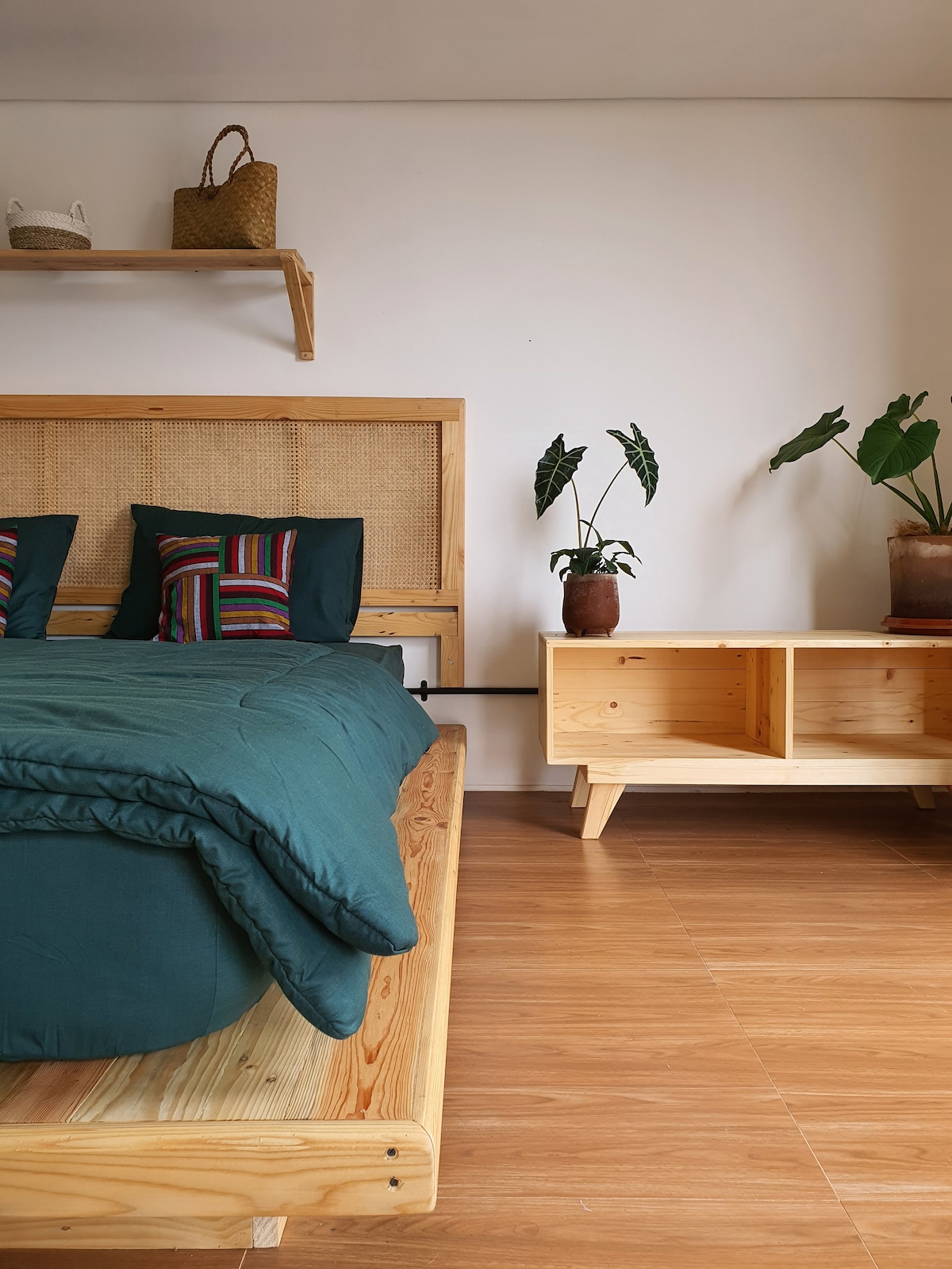 A serene bedroom features a wooden bed frame with a cozy green duvet, complemented by a striped pillow arrangement. Natural light enters from a window, illuminating the space. Potted plants add a touch of greenery, while a minimalist wooden table provides functional storage.