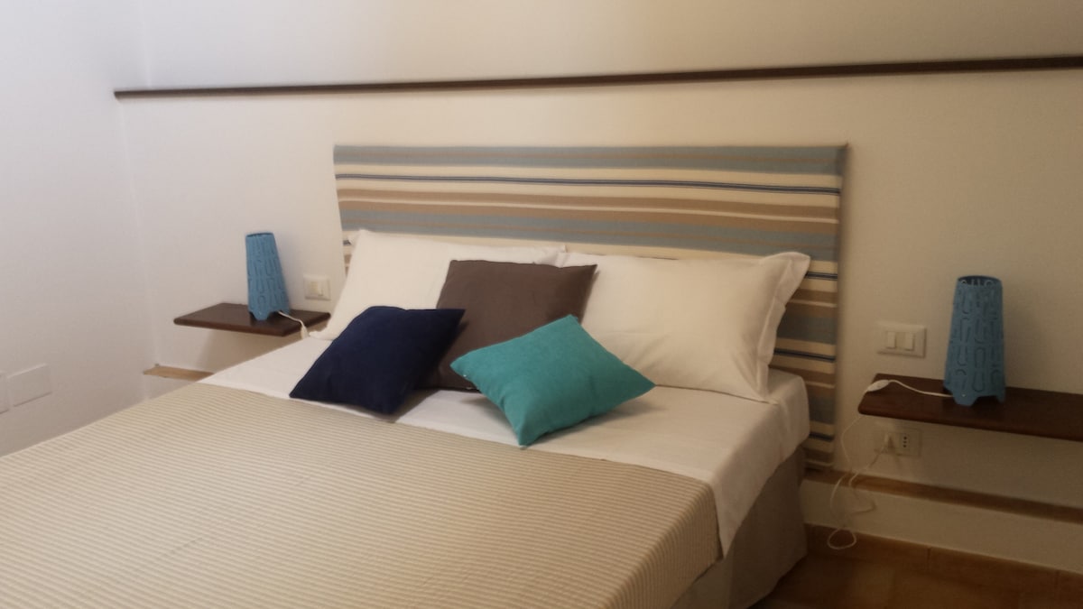 A serene bedroom features a spacious bed with a striped headboard, adorned with decorative pillows in blue and grey. Nightstands with blue lamps are positioned on either side, and warm light creates a calming environment.
