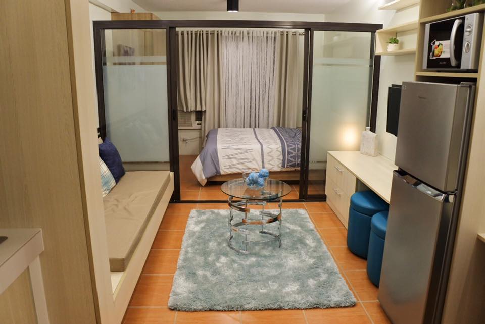 A modern studio layout features glass partitions separating the sleeping area from the living space. A cozy sofa lies adjacent to a low glass table, with a soft rug beneath. The modular kitchen is equipped with a refrigerator and microwave. Natural light enters through sheer curtains.