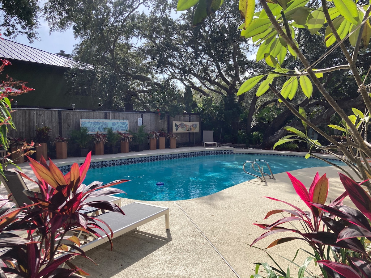 The image captures a sunny outdoor pool area surrounded by vibrant foliage. A spacious swimming pool is visible, accompanied by lounge chairs placed nearby. Colorful planters with tropical plants enhance the landscape, creating a relaxed atmosphere in the backyard.