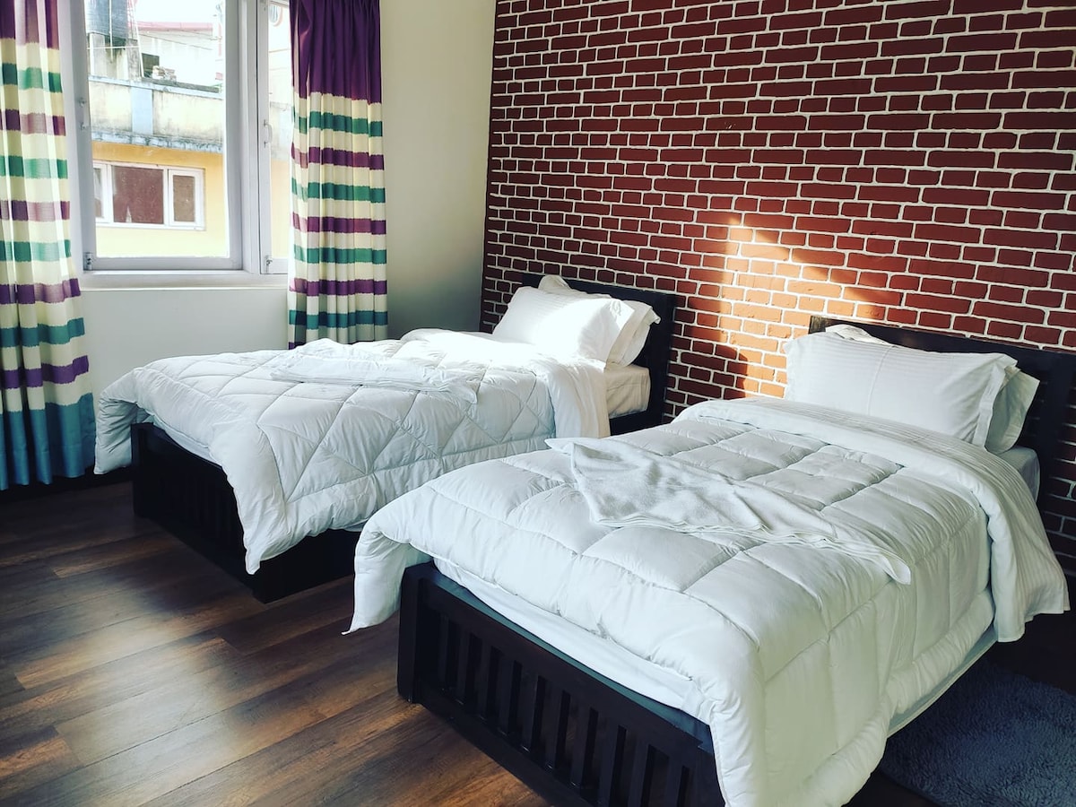 A bright room features two single beds, each covered with white bedding. A textured brick wall adds character, while large windows with striped curtains allow natural light to enhance the space. A simple rug lies on the wooden floor, providing comfort underfoot.
