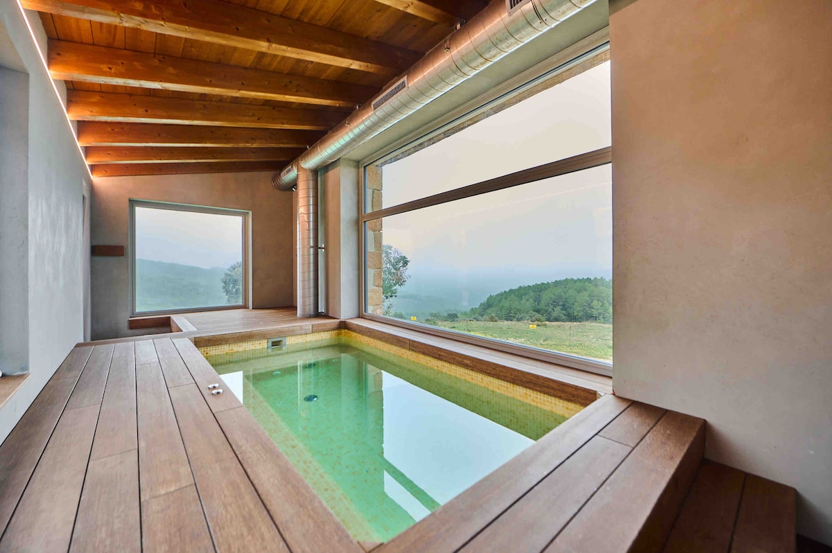 An indoor pool is framed by wooden decking, creating a relaxing area. Large windows provide views of the surrounding landscape, allowing natural light to fill the space. The ceiling features wooden beams, enhancing the architectural design.