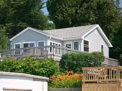 A cottage with light grey siding is set against a backdrop of greenery. It features a wraparound wooden deck with multiple windows that provide views of the surrounding landscape. The front yard displays vibrant flowers, enhancing the outdoor environment.