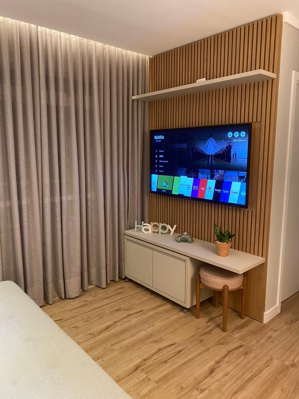 A modern entertainment area is displayed, featuring a large, wall-mounted television. Below, a sleek, white media console offers storage. Soft gray curtains frame the window, while a small potted plant adds a touch of greenery to the cozy space.
