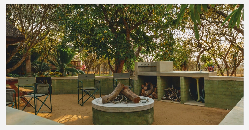 A fire pit is centrally located within a circular stone structure, surrounded by several folding chairs. A stone bench is adjacent, with stacked firewood neatly arranged nearby. Lush greenery and trees provide a natural backdrop, creating a serene outdoor setting.