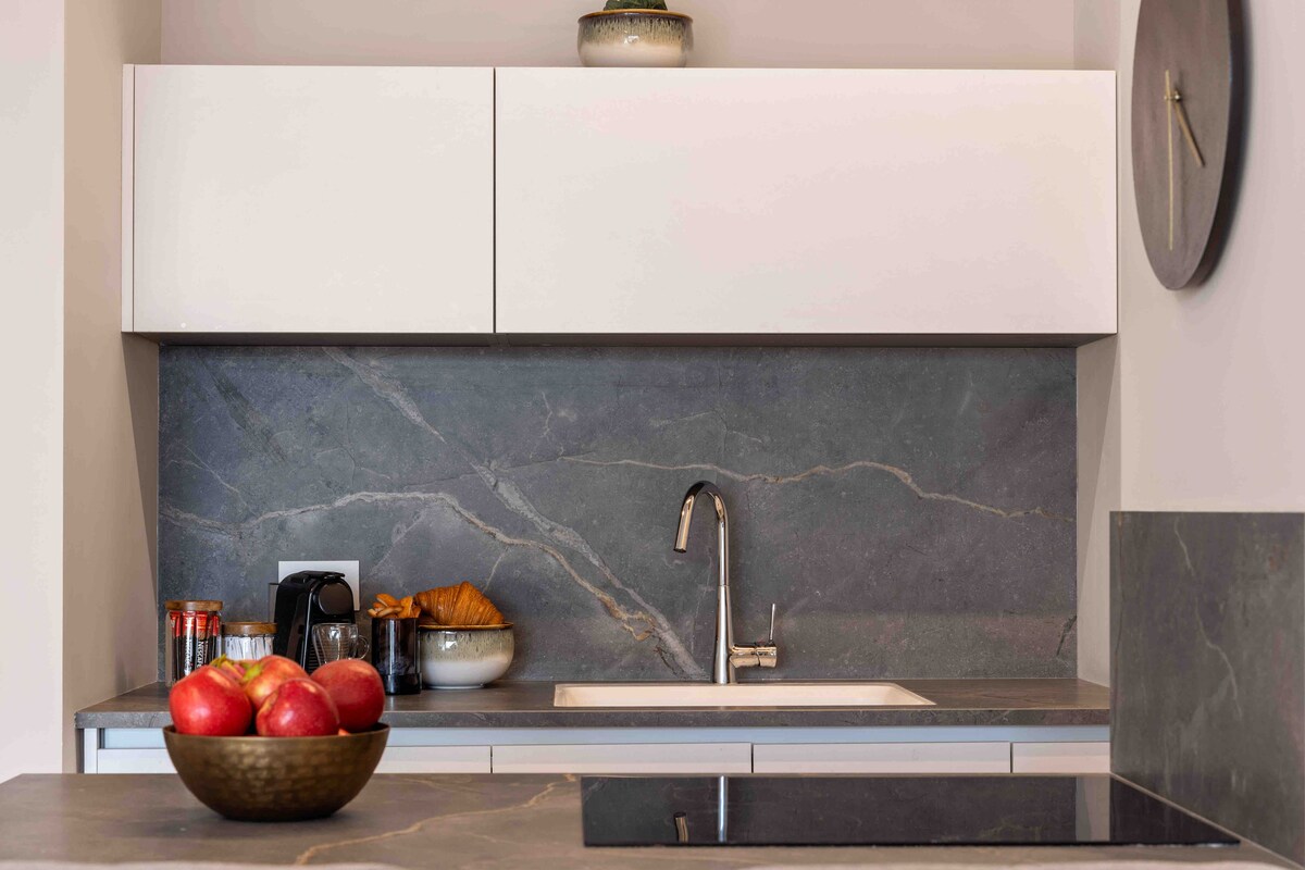 A modern kitchen is showcased with sleek cabinetry and a dark stone backsplash. A sink and stovetop are integrated into the countertop. A bowl of apples and a plate of pastries are placed on display, along with a Nespresso coffee machine and a jar of supplies.