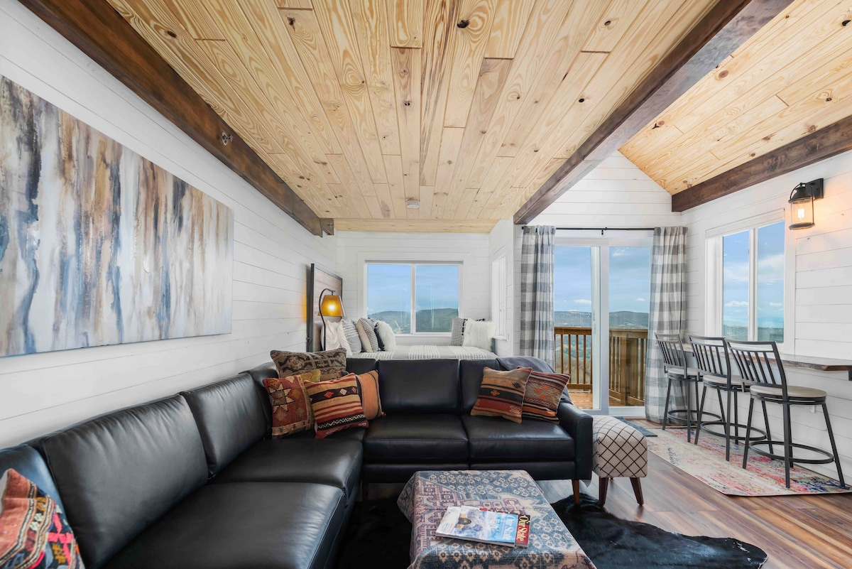 A spacious living area features a high vaulted ceiling with wooden beams. A large leather sectional sofa is arranged around a patterned coffee table. Expansive windows frame scenic views of the outdoors, while a dining area with stools is positioned near the windows. Natural light brightens the space.