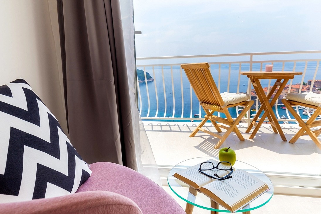 A comfortable seating area is visible, featuring a soft pink chair and a glass table. An open book rests on the table, accompanied by reading glasses and a green apple. Beyond the glass door, a small balcony is seen with two wooden chairs and a table overlooking the sea.