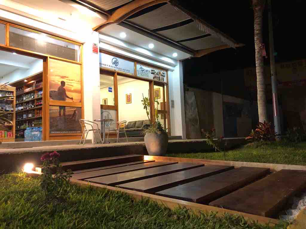 An inviting entrance is shown at night, featuring wooden pathways leading to a modern building. Soft lighting illuminates the area and a small outdoor seating arrangement with chairs and a table is visible. Lush green grass and flower beds add to the surroundings.