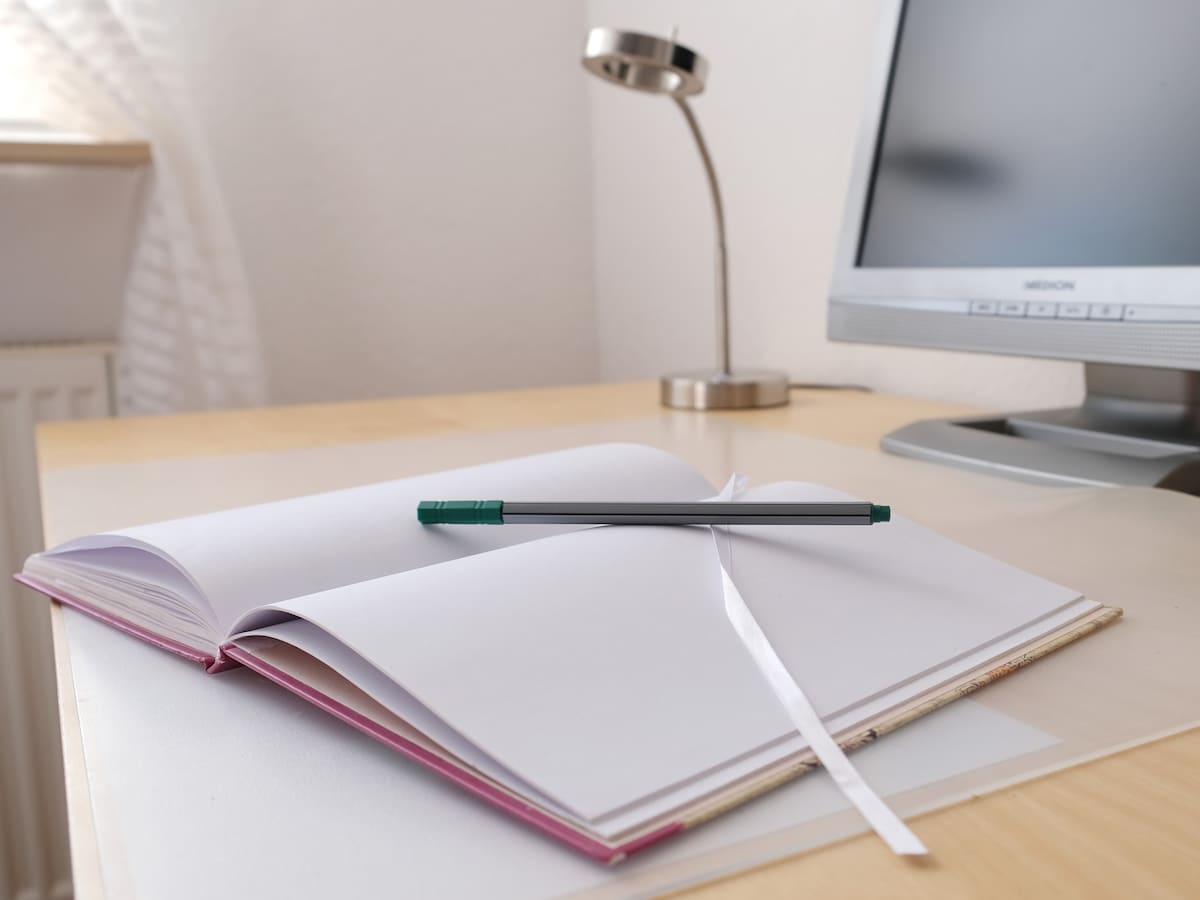 A work desk is seen with an open notebook featuring blank pages, accompanied by a green pen resting across it. A desk lamp and a computer monitor are visible in the background, contributing to a functional workspace atmosphere.