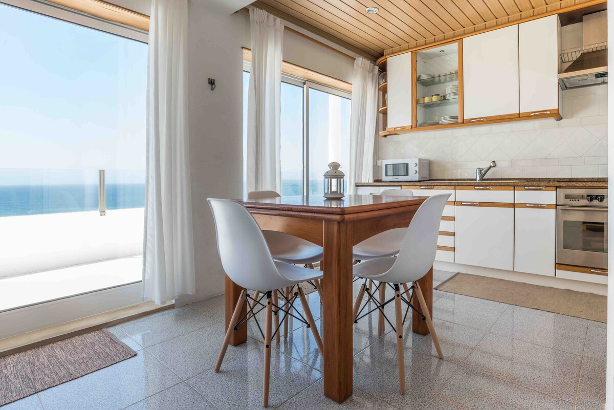 A bright kitchen area features a wooden dining table surrounded by four white chairs. Large windows provide unobstructed ocean views, while cabinetry offers ample storage. A microwave and oven are included in the well-equipped kitchen, and soft curtains allow natural light to fill the space.