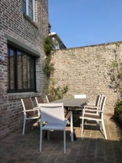 A private outdoor patio area is displayed, featuring a large table surrounded by six light-colored chairs. The brick wall provides a textured backdrop, with greenery subtly enhancing the space. Clear blue skies can be seen above, creating a bright and inviting atmosphere.