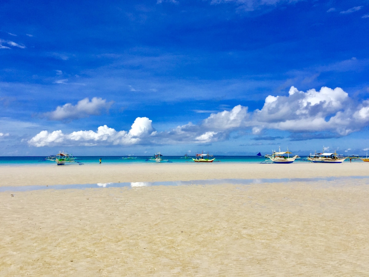 A serene beach scene showcases a vast stretch of white sand under a bright blue sky. The clear water reflects the clouds and boats in the distance, creating a tranquil coastal landscape. The unobstructed view emphasizes the peacefulness of the beachfront.
