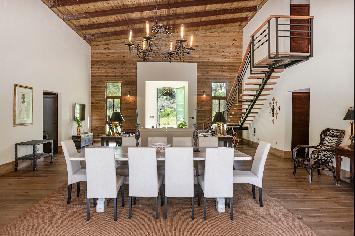 A spacious dining area features a large table surrounded by six upholstered chairs, complemented by warm wooden accents. Natural light fills the room through large windows, and a staircase with a wooden railing leads to the upper level. Decorative elements include a chandelier and framed artwork.