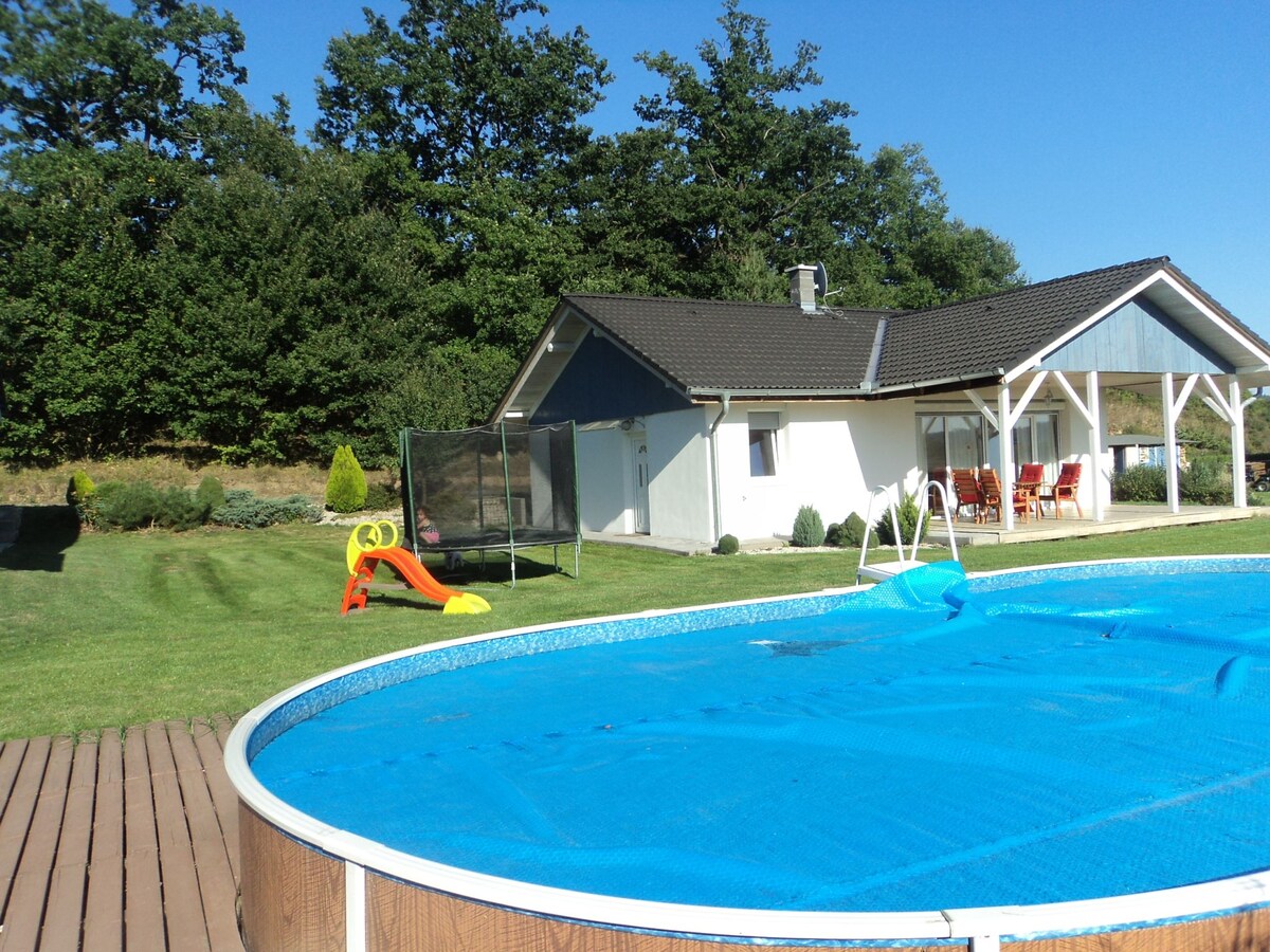 A swimming pool is surrounded by a wooden deck, with a safety cover in place. In the background, a bungalow features a covered terrace, outdoor seating, and a large trampoline. Lush greenery complements the peaceful landscape, providing a spacious area for relaxation.