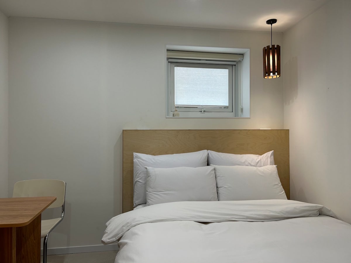 A bedroom features a spacious bed with white linens against a light wooden headboard. A small wooden table and chair are located beside the bed. Natural light enters through a window, and a pendant light adds soft illumination to the space.