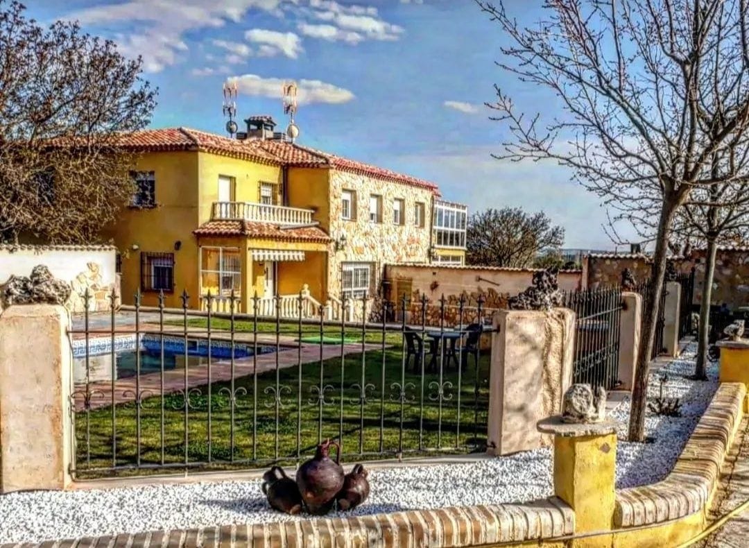 A charming multi-story home is surrounded by a well-maintained garden featuring a fenced area and a clear blue swimming pool. Outdoor seating is arranged under the sun, with a few trees providing shade. Decorative elements enhance the inviting exterior of the property.