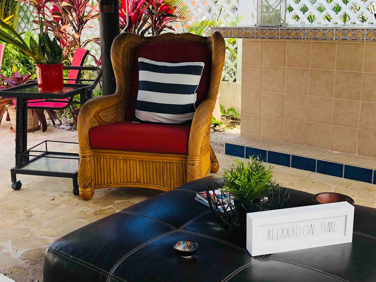 A cozy outdoor seating area is presented, featuring a rattan chair with a red cushion and striped throw pillow. A low coffee table displays decorative items and a small plant. Lush greenery surrounds the space, contributing to a relaxed atmosphere.