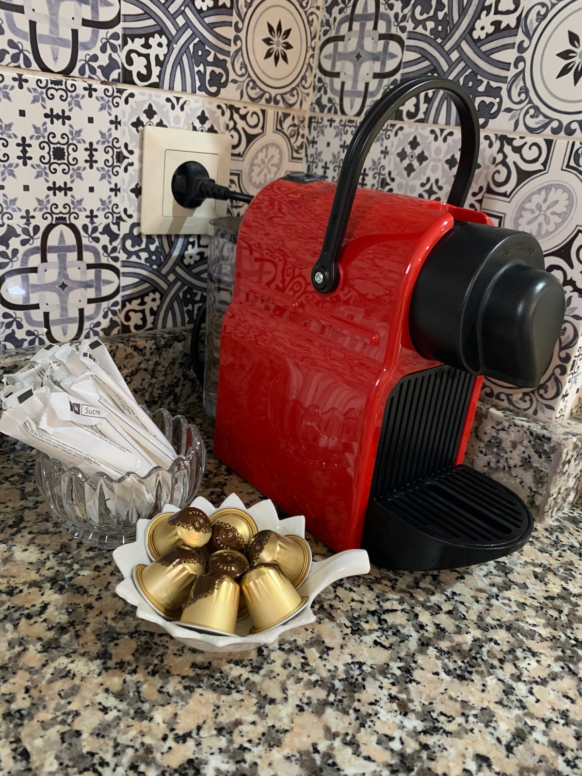 A modern coffee machine in vibrant red sits on a speckled granite countertop. Next to it, a bowl holds assorted coffee capsules. A small dish with coffee stirrers is also present, adding convenience for guests. The colorful tile backsplash enhances the kitchen's inviting appearance.