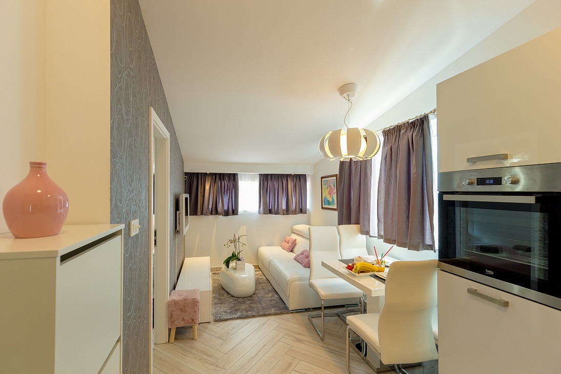 A modern living area with a sectional sofa is complemented by a light-colored dining table and four chairs. Natural light filters through large windows with dark curtains. A kitchenette is visible, featuring sleek appliances and minimalist decor.