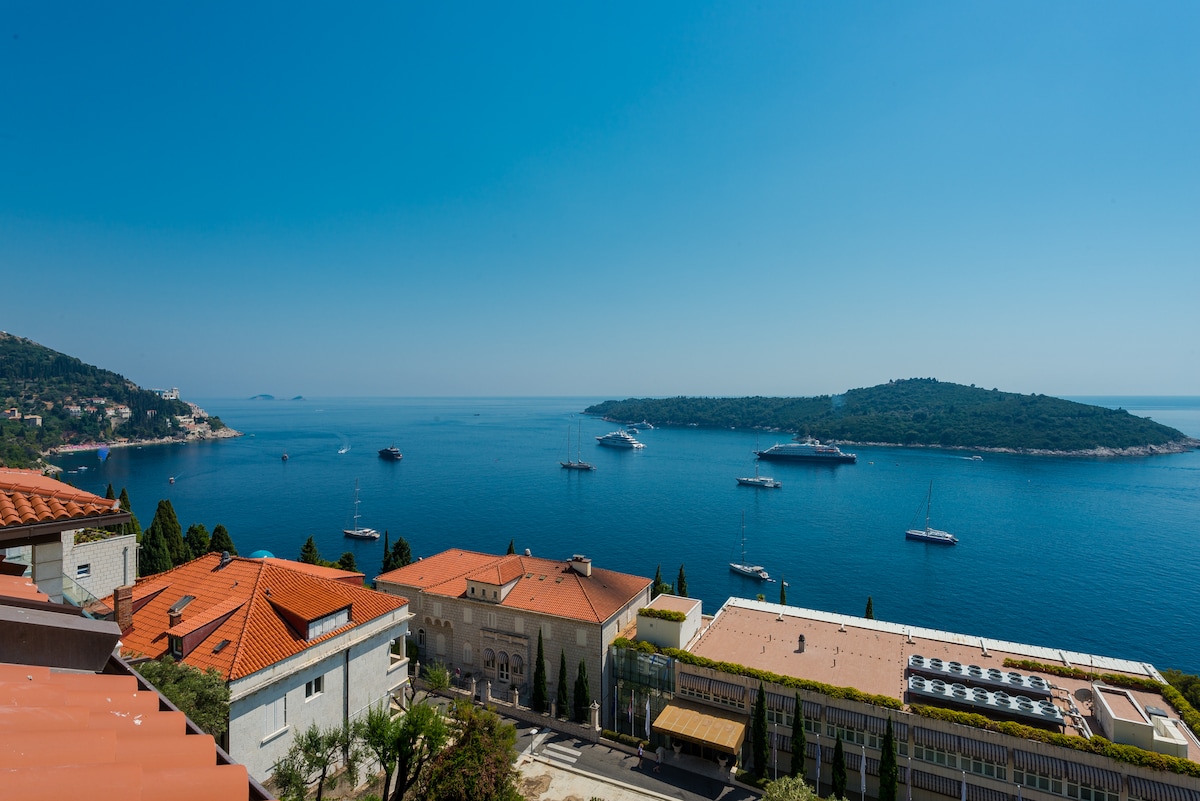 A panoramic view is presented, showcasing the tranquil blue sea dotted with boats, with the island of Lokrum visible in the distance. The landscape features terracotta rooftops and lush greenery, creating a serene coastal atmosphere. A clear sky enhances the beauty of the setting.