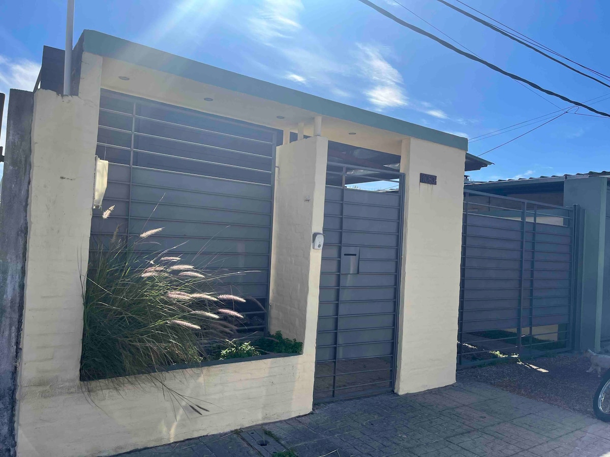 A contemporary exterior is displayed with a combination of light-colored walls and gray accents. A secure gate opens to a small entrance pathway, surrounded by minimal landscaping, featuring tall grasses. The clear blue sky serves as a backdrop, enhancing the modern design.