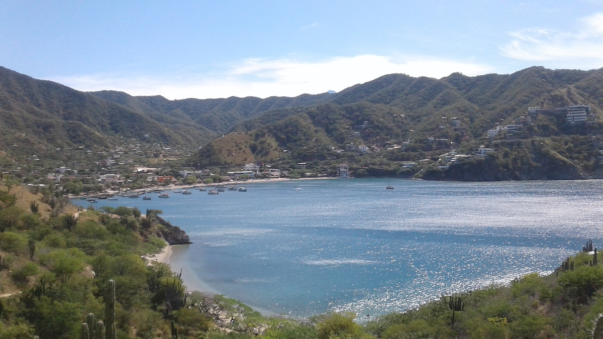A serene bay is depicted, surrounded by lush green hills and mountains. Several boats are gently floating on the water, which reflects the sunlight creating a shimmering effect. The landscape features a mix of coastal vegetation and distant residential areas.