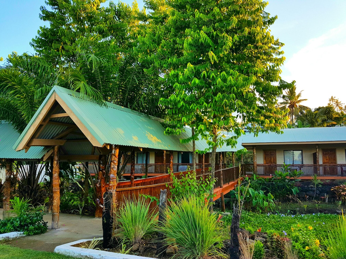 A charming building features a rustic design with a sloped roof and wooden accents. Lush greenery surrounds the structure, including tall trees and tropical plants. A path leads to the entrance, offering accessibility and a welcoming ambiance for families.