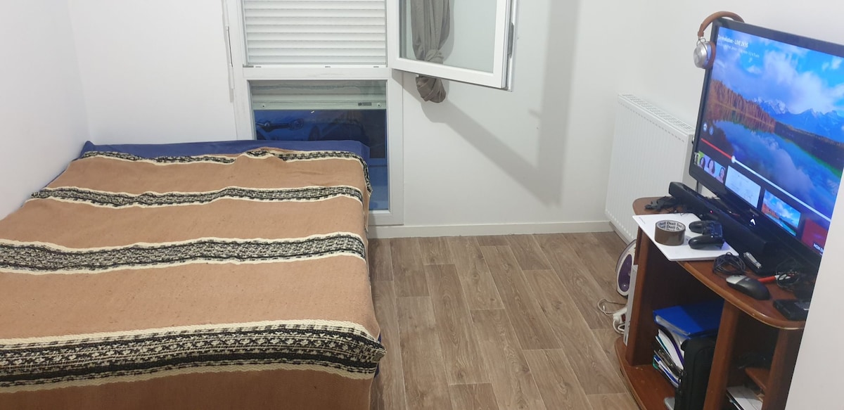 A cozy sleeping area is furnished with a spacious bed covered by a patterned blanket. A television is positioned on a wooden stand beside the bed, and light enters through an open window, which is dressed with light-colored blinds.