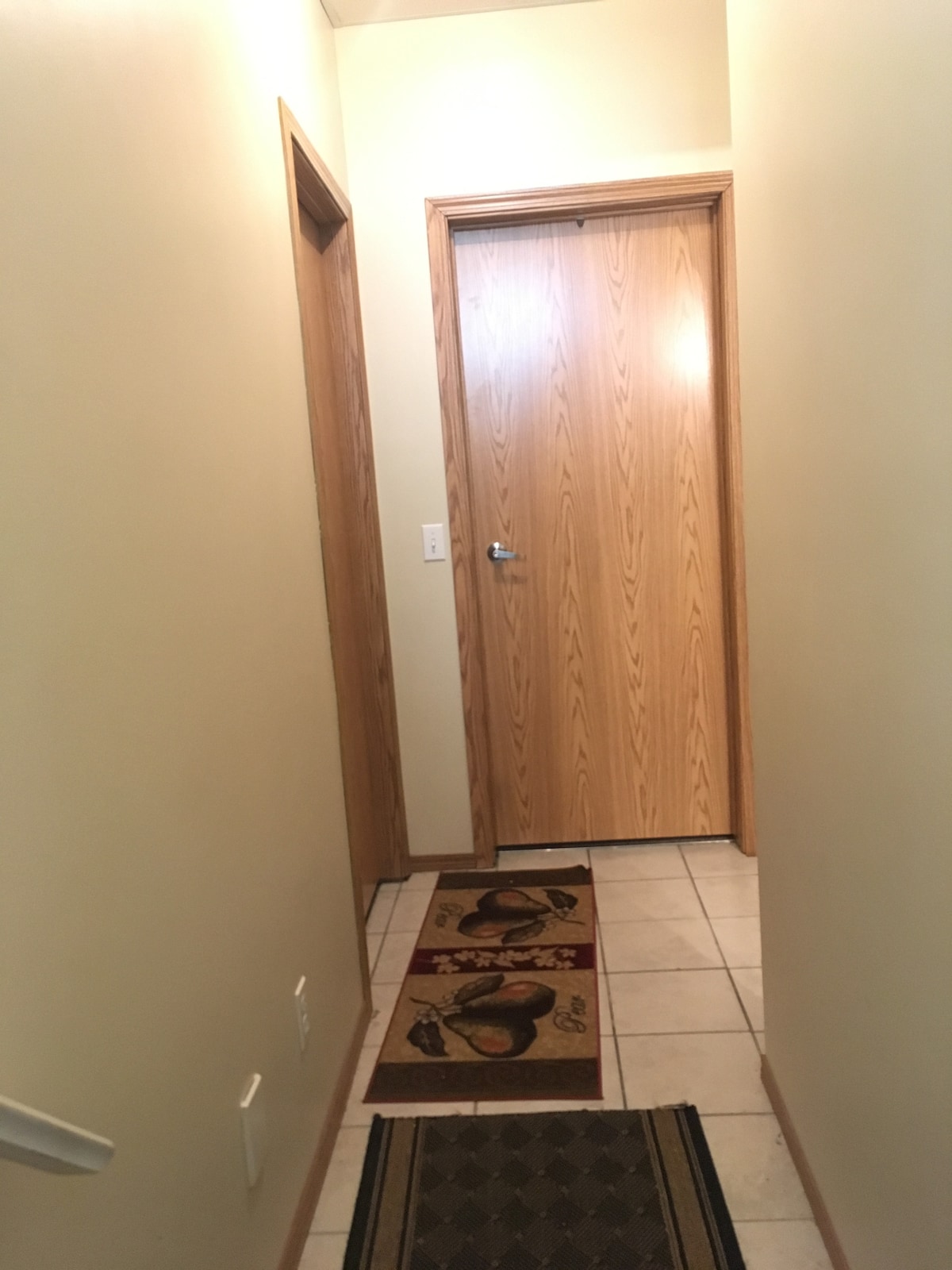 A narrow hallway is presented with warm-toned walls and tiled flooring. A patterned area rug lies in the center, while wooden doors are visible on either side. Soft lighting enhances the welcoming atmosphere of the space, leading towards a closed door at the end.
