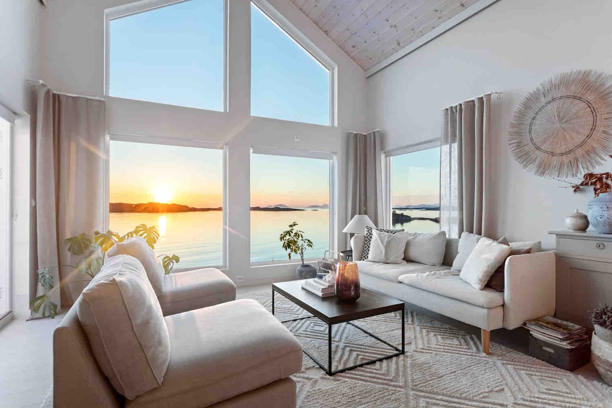 A spacious living area is illuminated by large windows, offering a view of the sunset reflecting on the water. Light-colored sofas and a coffee table create a comfortable setting, while soft curtains frame the windows, enhancing the serene ambiance.