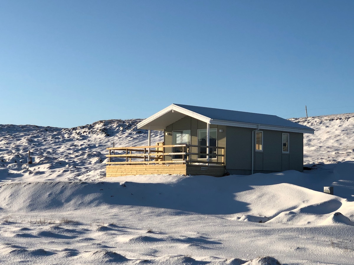 A cottage sits against a backdrop of snow-covered terrain under a clear blue sky. A wooden deck extends from the cottage, providing outdoor space, while the structure features a light-coloured exterior with large windows for natural light.