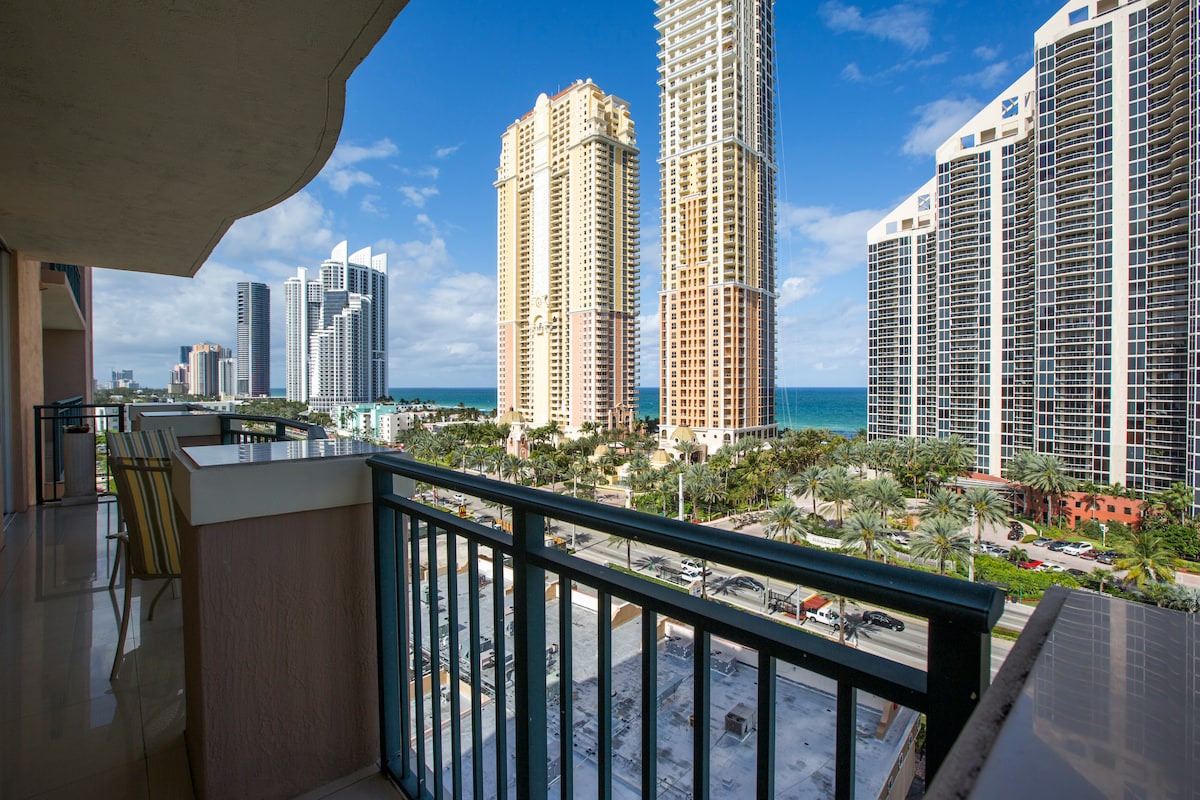 A spacious balcony offers a view of the city skyline and the ocean in the distance. Tall buildings rise around the area, while palm trees line the streets below, creating a lively atmosphere. The clear blue sky enhances the urban setting.