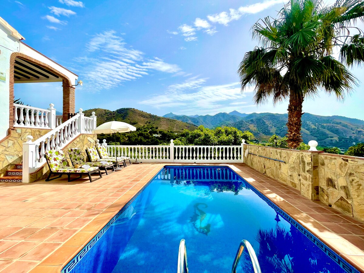 A private pool is set against a backdrop of mountains, complemented by a palm tree and sun loungers. A spacious terrace features a vibrant blue sky, casting natural light over the area. The pool's steps provide easy access for guests.