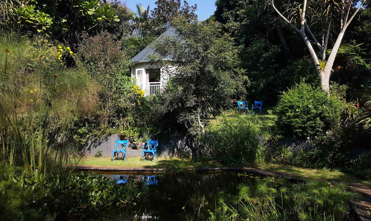 A serene garden setting includes a small cottage with a gabled roof, surrounded by lush greenery and flowering plants. A reflective pond is visible in the foreground, with vibrant blue chairs arranged in the garden, offering a peaceful outdoor space.