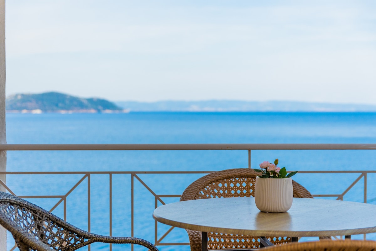 A small round table is set with a potted plant, surrounded by two woven chairs on a spacious veranda. The view reveals the open sea and a distant island against a clear blue sky, enhancing the tranquil setting.