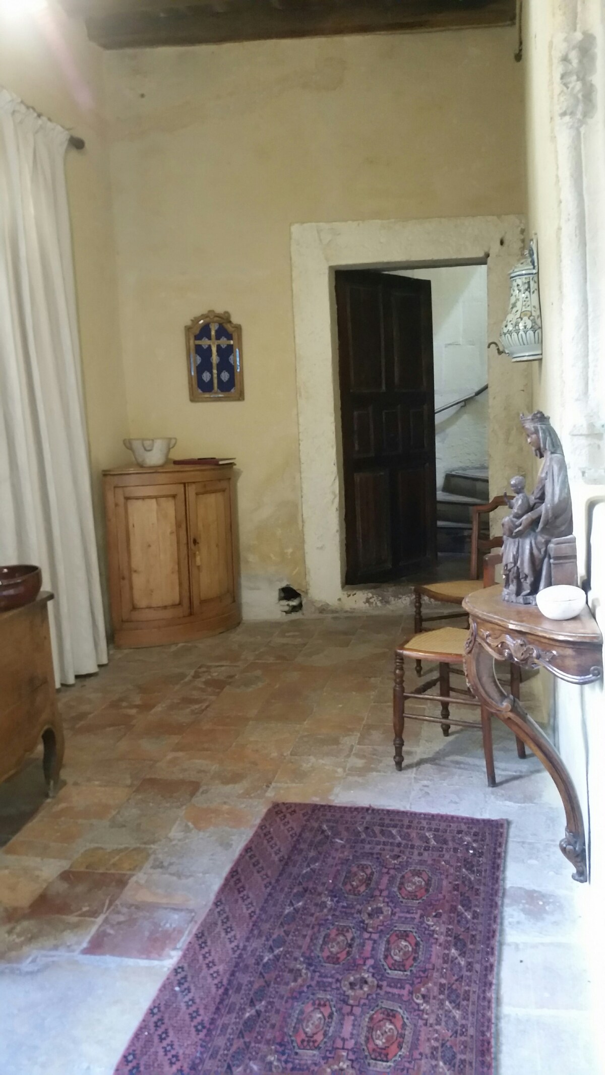The entrance hall showcases rustic stone flooring with a colorful area rug. Light beige walls are complemented by wooden furniture, including a cabinet and a small decorative table. A staircase leads to the upper level, while a statue and wall decor add character to the space.