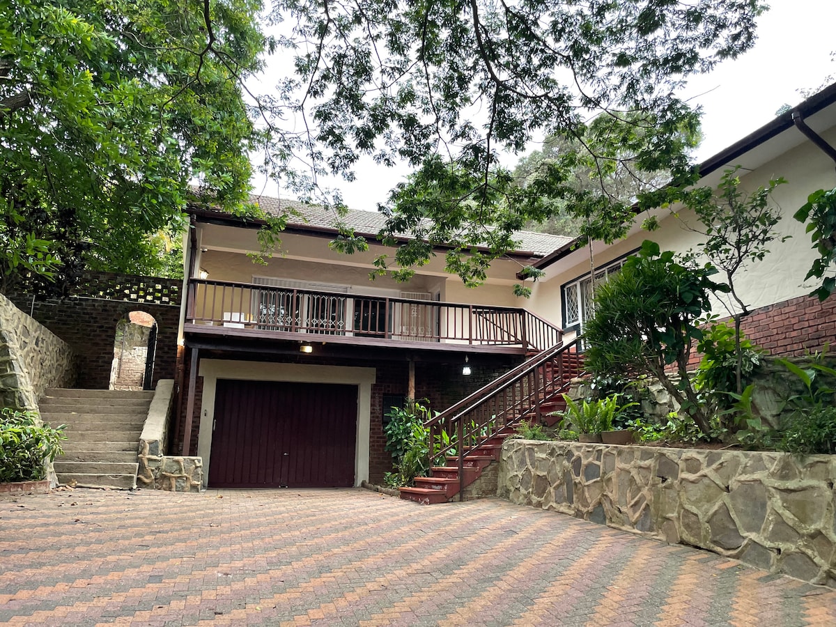 The exterior of the house is showcased, featuring a stone-paved driveway leading to a lock-up garage. A welcoming entrance is framed by greenery, and a staircase ascends to a covered porch with railing. Lush plants add to the natural ambiance surrounding the property.