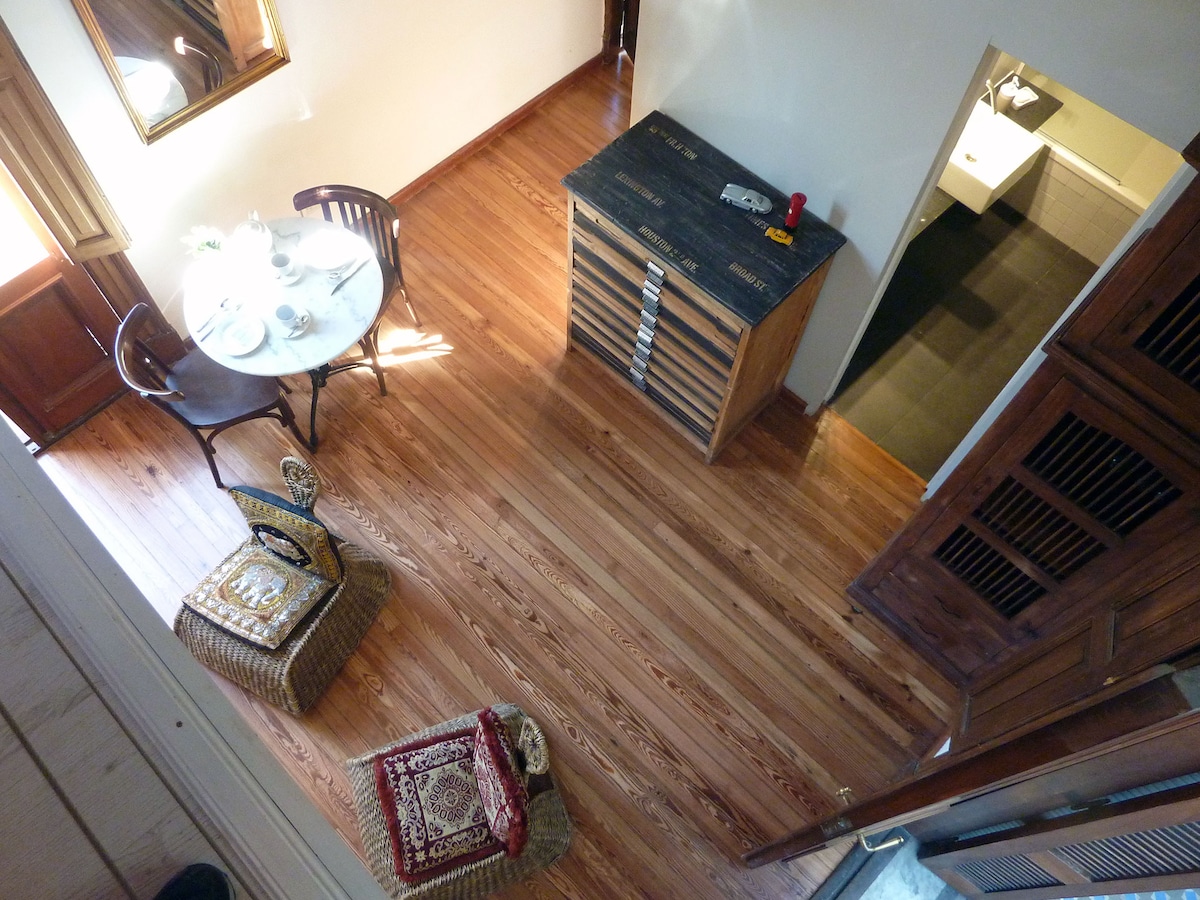 A wooden floor with a warm tone is highlighted beneath a small dining table set for two, featuring light dishes. Nearby, woven seating is arranged beside a chest with a rustic finish. Natural light filters through nearby windows, enhancing the inviting space.