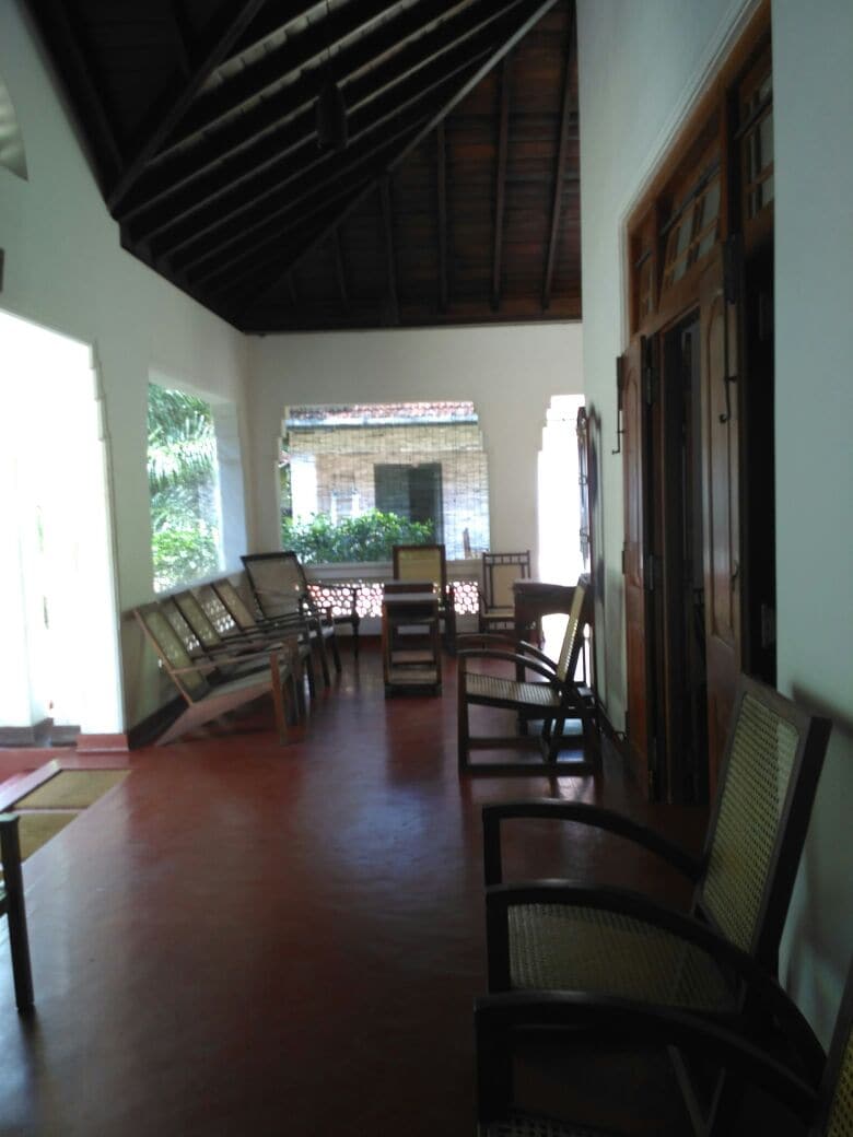 A spacious porch features multiple wooden chairs arranged along the walls, providing a comfortable area for relaxation and conversation. Large windows allow natural light to enter, revealing greenery from the outside. The high wooden ceiling enhances the airy feel of the space.