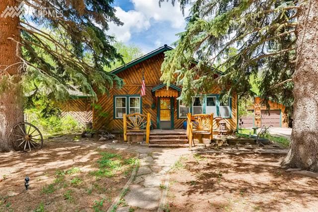 A charming cabin, framed by tall evergreen trees, features a prominent entryway with rustic wooden steps. A welcoming porch is equipped with comfortable seating, surrounded by natural landscaping. The structure showcases a traditional wood exterior, highlighted by a colorful doorway, under a blue sky with fluffy clouds.