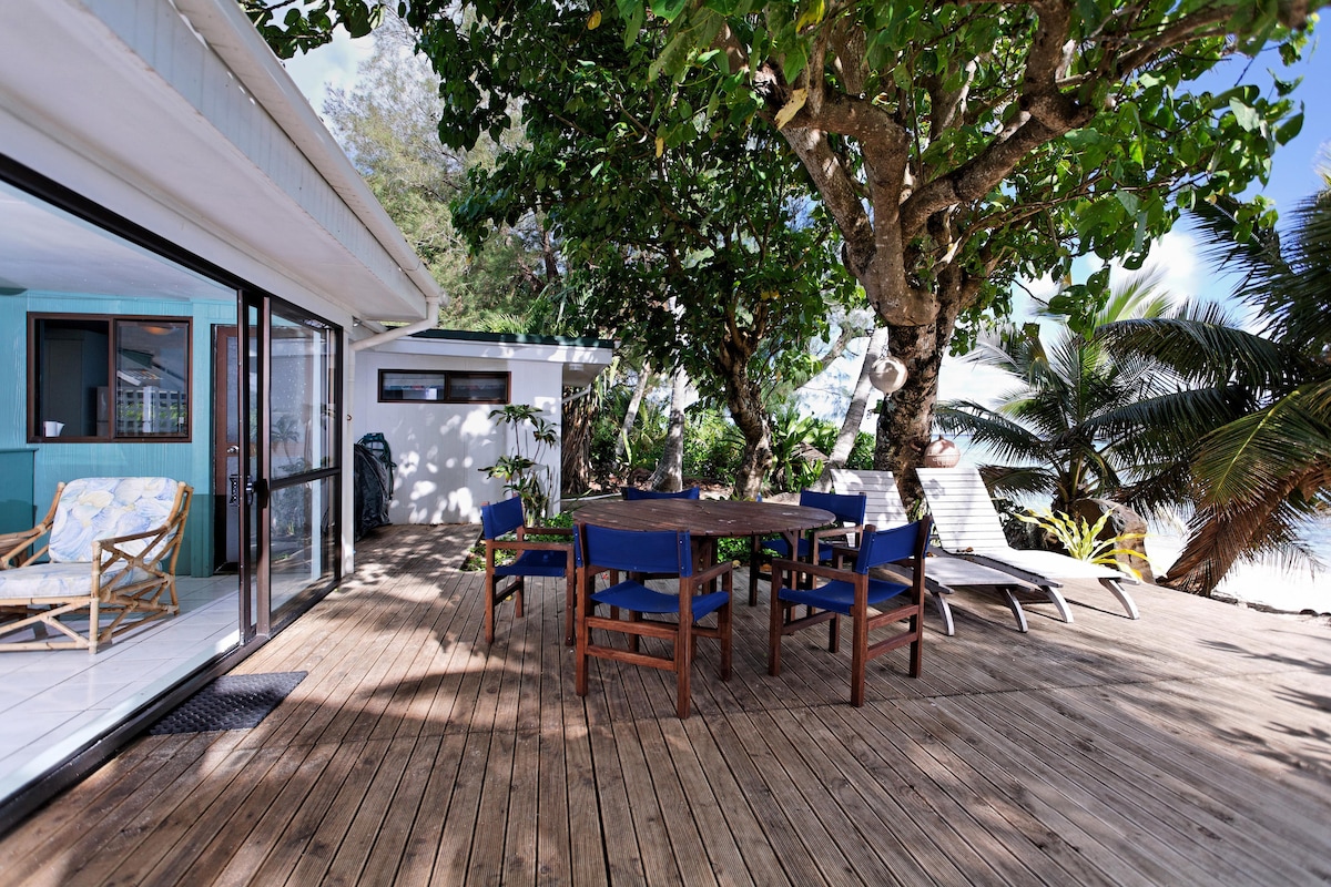 An outdoor deck area featuring a round wooden table surrounded by blue chairs, providing a space for dining or relaxation. The deck is shaded by lush trees and allows easy access to the interior living space through sliding glass doors.