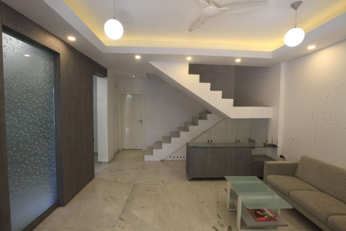 An open living area features a light grey sofa positioned next to a glass coffee table. A modern staircase ascends in the background, leading to the upper floor. Soft lighting highlights the clean lines of the space, while a decorative wall adds texture to the setting.