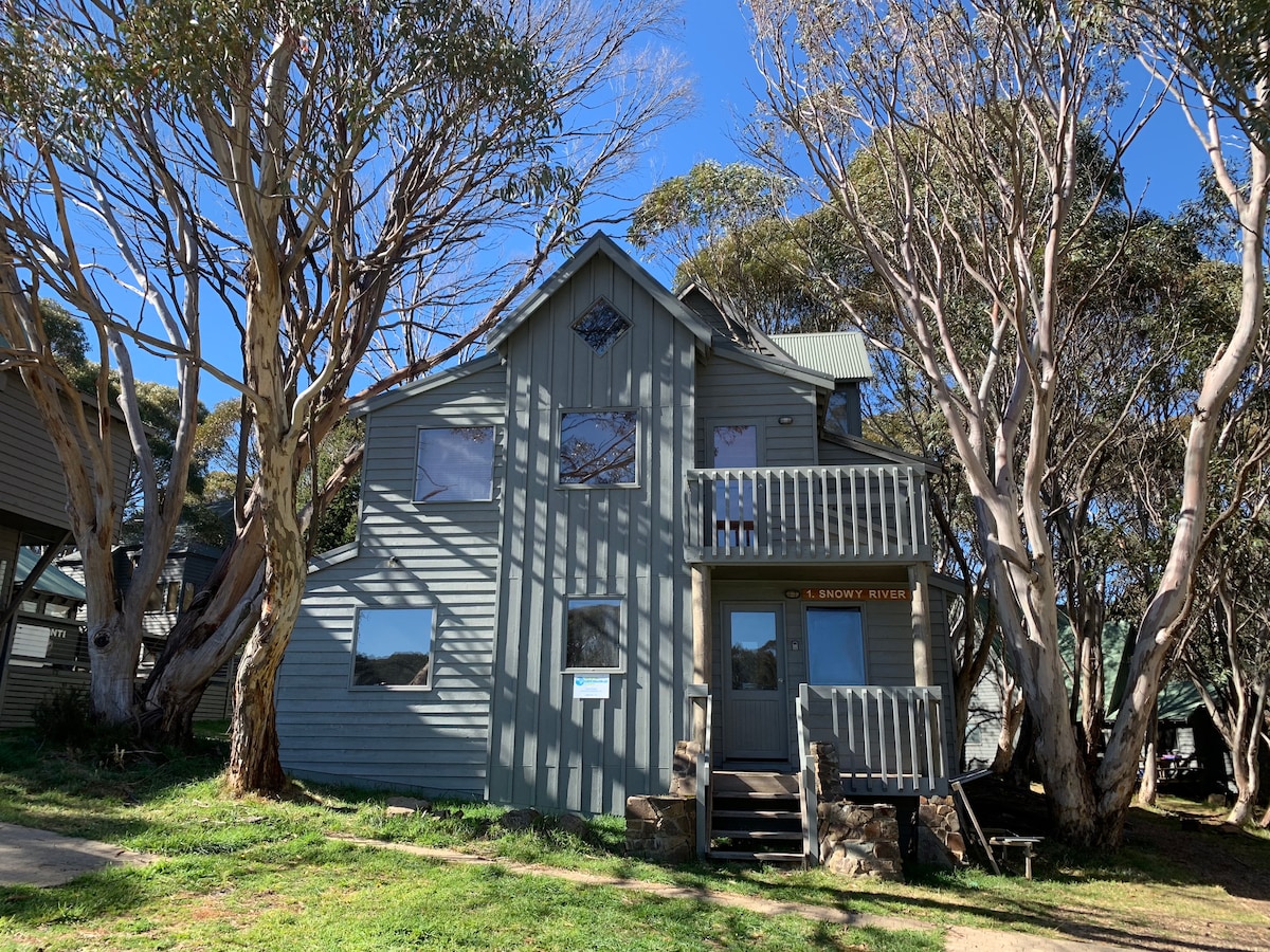 A quaint, multi-level structure is framed by tall, slender trees and green grass. The building features a mix of large windows and a welcoming entrance, with a small porch and decorative detailing on the upper section. A clear blue sky complements the exterior.