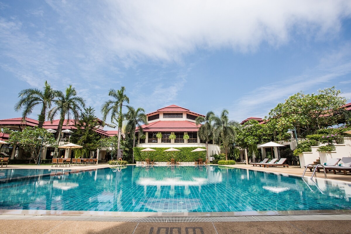 A large swimming pool is surrounded by palm trees and lounge chairs, inviting guests to relax. Resort-style buildings in the background are framed by lush greenery, with outdoor seating areas visible beneath shaded umbrellas.